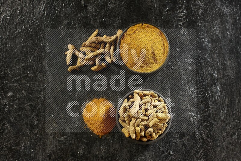 2 pottery black bowls, one full of turmeric powder and the other full of dried turmeric whole fingers on a textured black flooring