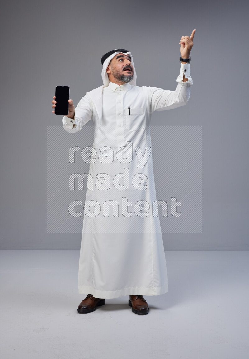 Saudi man Wearing Thob and white Shomag standing showing phone to camera on Gray background