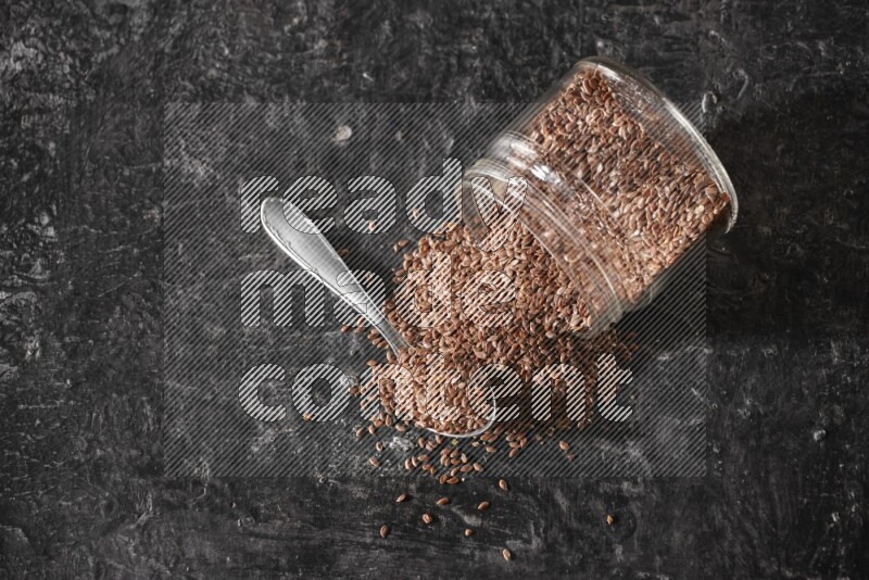 A glass jar full of flaxseeds flipped and seeds spread out with a metal spoon full of the seeds on a textured black flooring