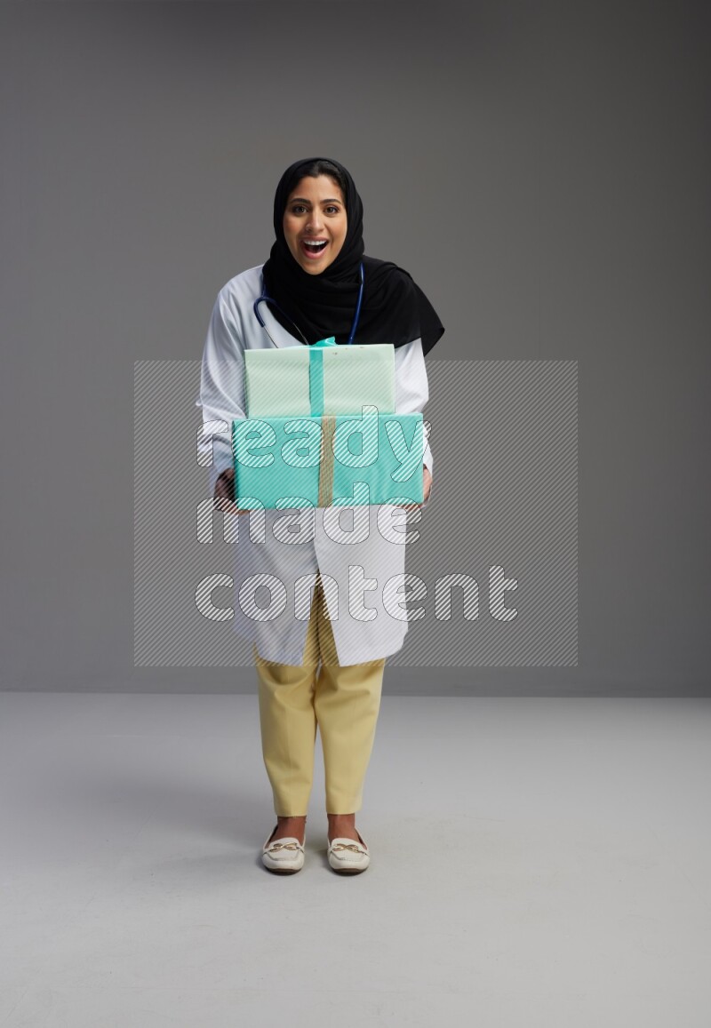 Saudi woman wearing lab coat with stethoscope standing holding gift box on Gray background