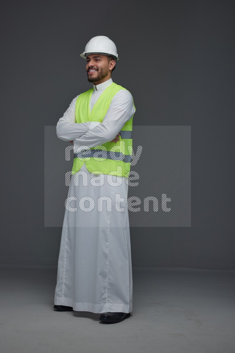 A Saudi man wearing Thobe with a yellow safety vest and white helmet standing and crossing his hands eye level on a gray background