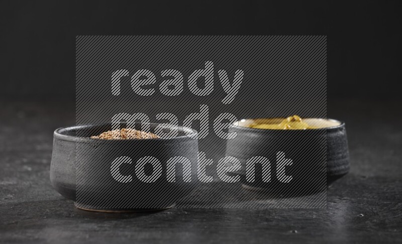 2 black pottery bowls full of mustard seeds and mustard paste on black flooring
