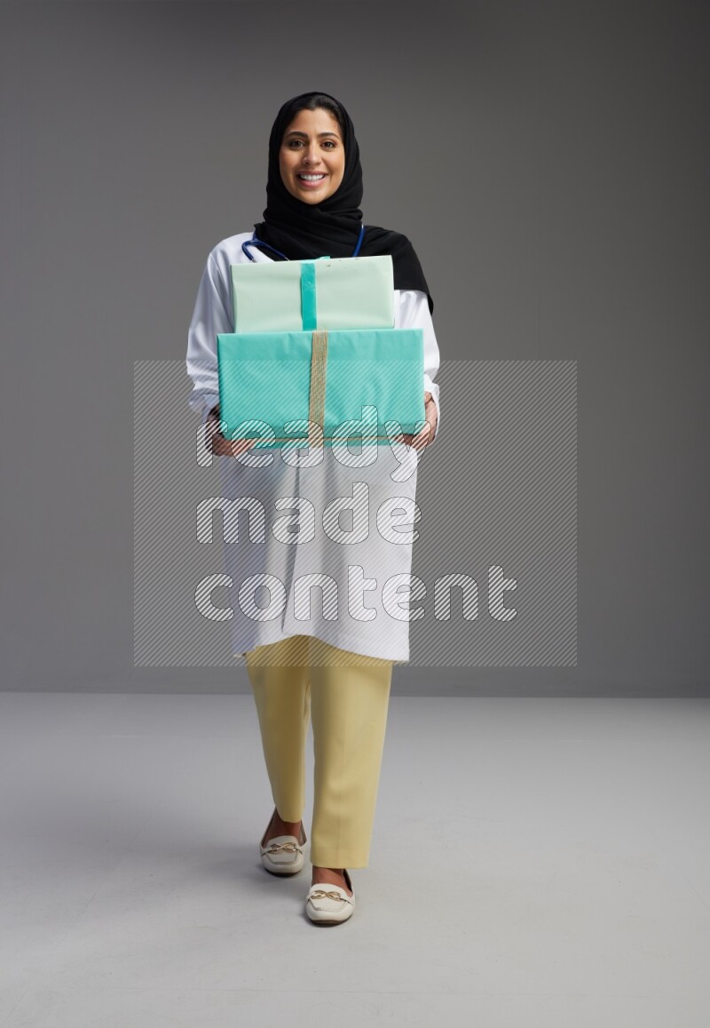 Saudi woman wearing lab coat with stethoscope standing holding gift box on Gray background