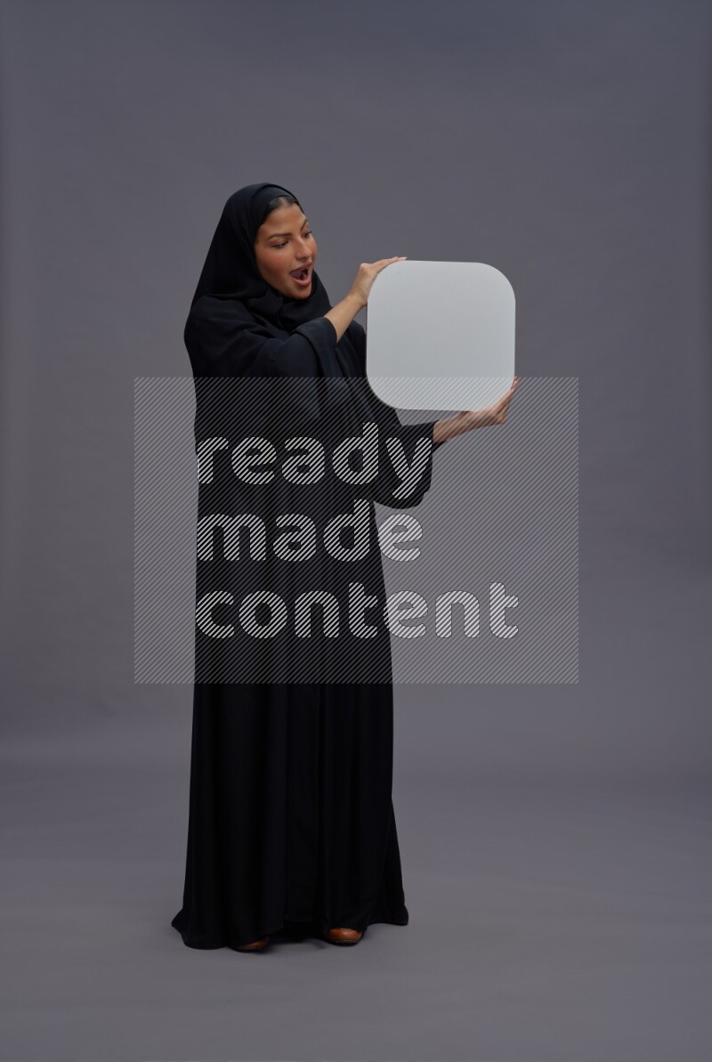 Saudi woman wearing Abaya standing holding social media sign on gray background