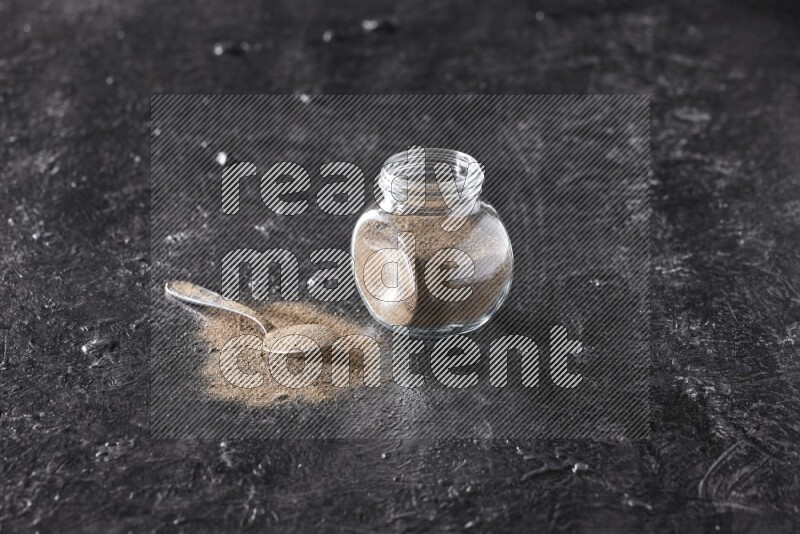 A glass spice jar full of black pepper powder and a metal spoon full of powder on textured black flooring