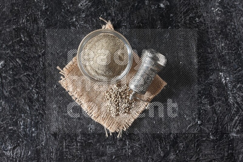 A glass bowl full of white pepper powder with white pepper beads on a burlap piece of fabric and a metal grinder on textured black flooring