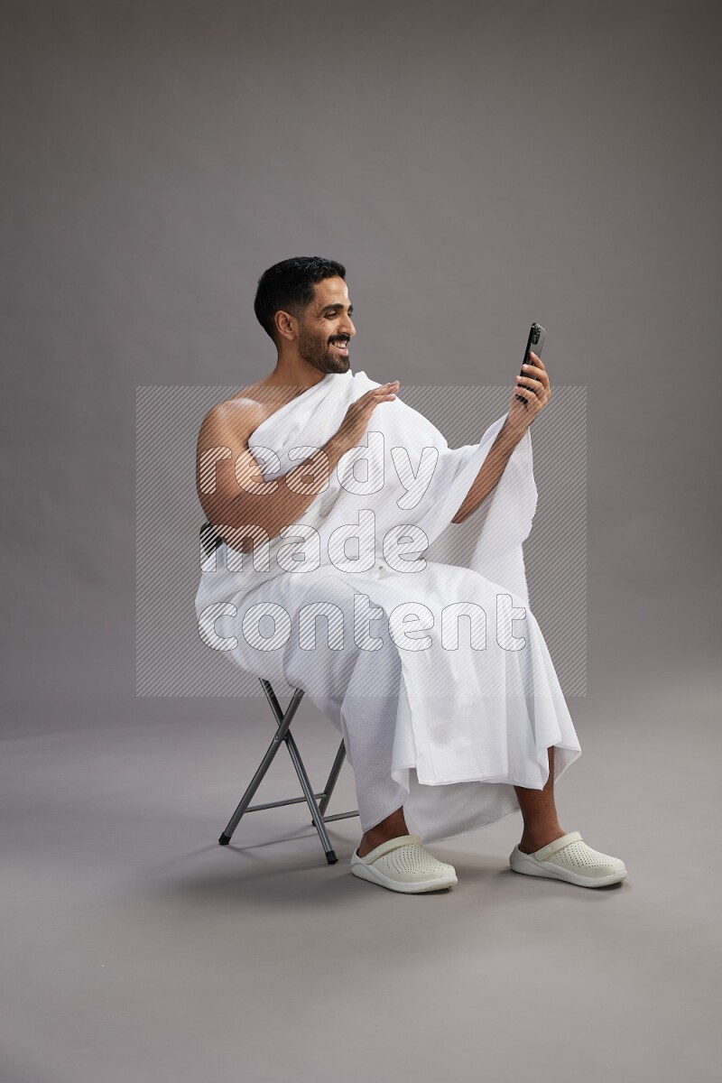 A man wearing Ehram sitting on chair taking selfie on gray background