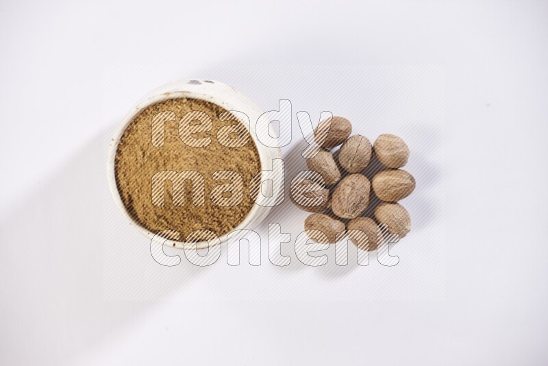 A beige pottery bowl full of nutmeg powder with whole seeds beside it on a white flooring