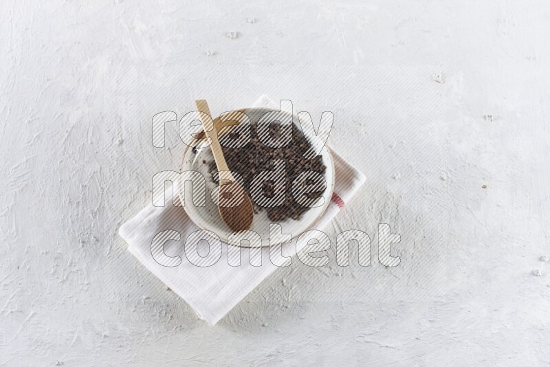 A Pottery plate full of whole cloves and a wooden spoon full of cloves powder in it on a textured white background