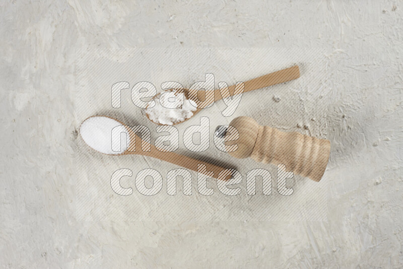 2 wooden spoons filled with fine and coarse salt with wooden grinder beside them on white background