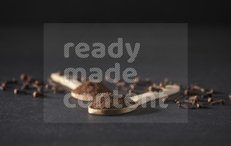 2 wooden spoons full of cloves powder with spreaded whole cloves on a black flooring