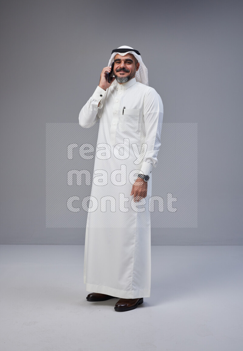 Saudi man Wearing Thob and white Shomag standing talking on phone on Gray background