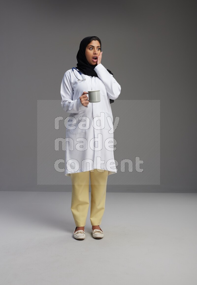Saudi woman wearing lab coat with stethoscope standing holding mug on Gray background