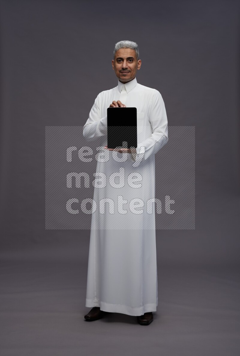 Saudi man wearing thob standing showing tablet to camera on gray background