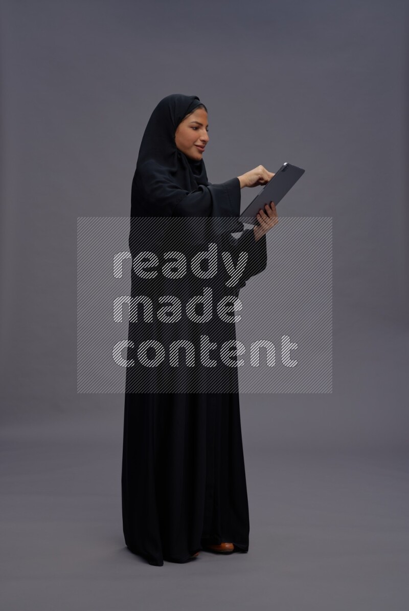 Saudi woman wearing Abaya standing working on tablet on gray background