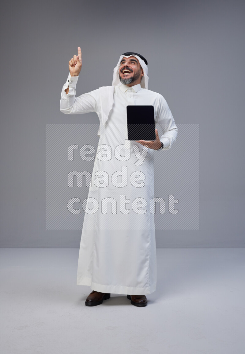 Saudi man Wearing Thob and white Shomag standing showing tablet to camera on Gray background
