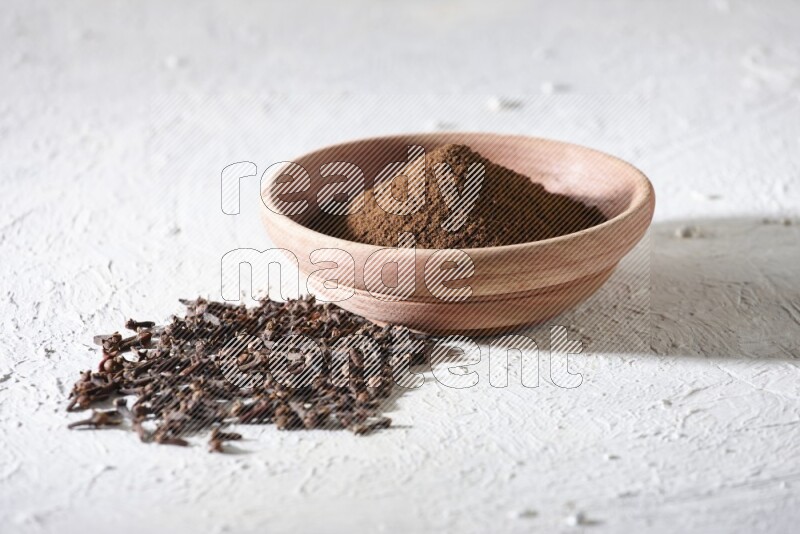A wooden bowl full of cloves powder with whole cloves beside it on a textured white flooring