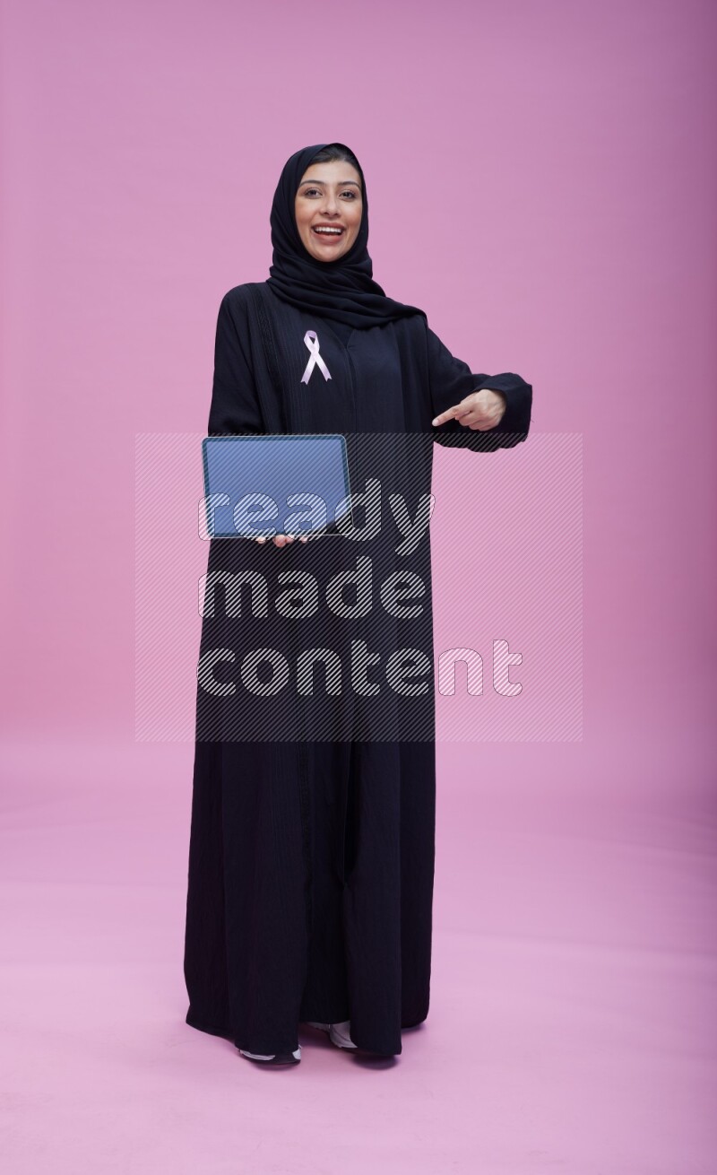 Saudi woman wearing pink ribbon on Abaya standing showing a tablet to camera on pink background