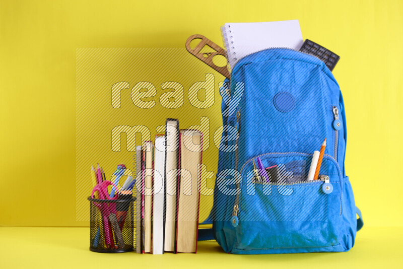 A school bag with assorted school supplies in and beside it on yellow background