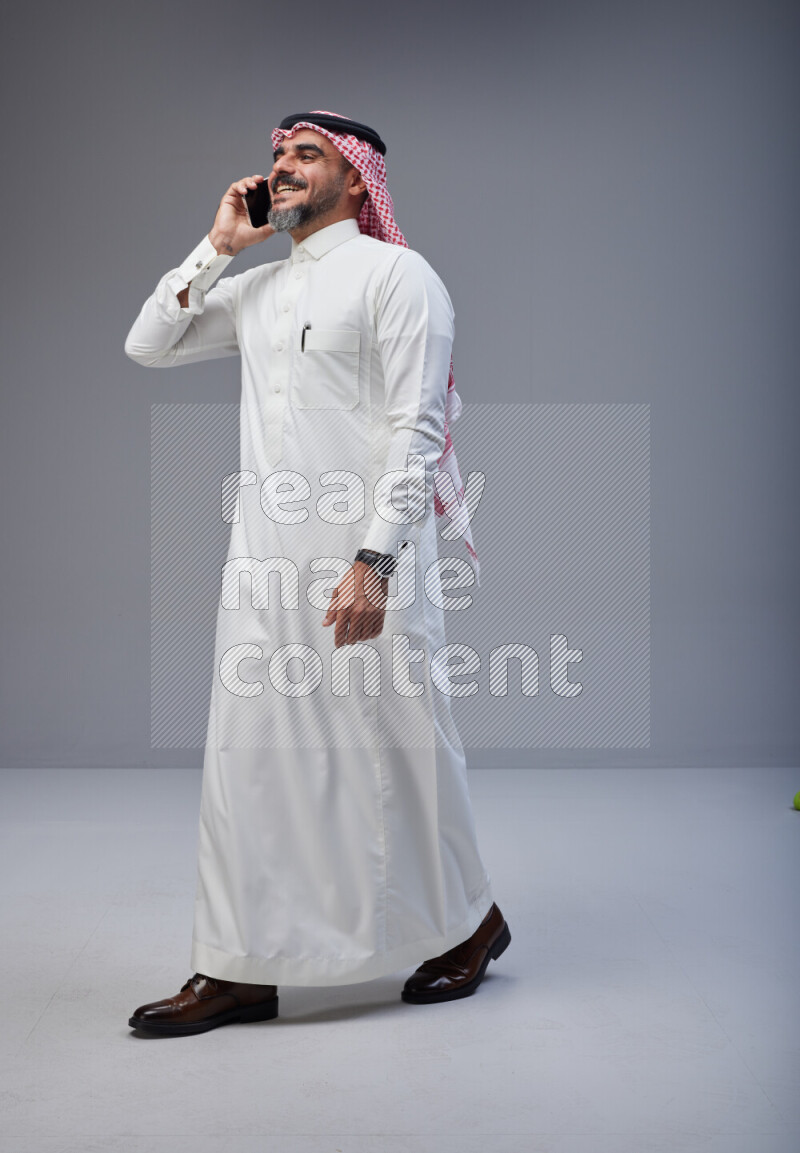 Saudi man Wearing Thob and red Shomag standing talking on phone on Gray background