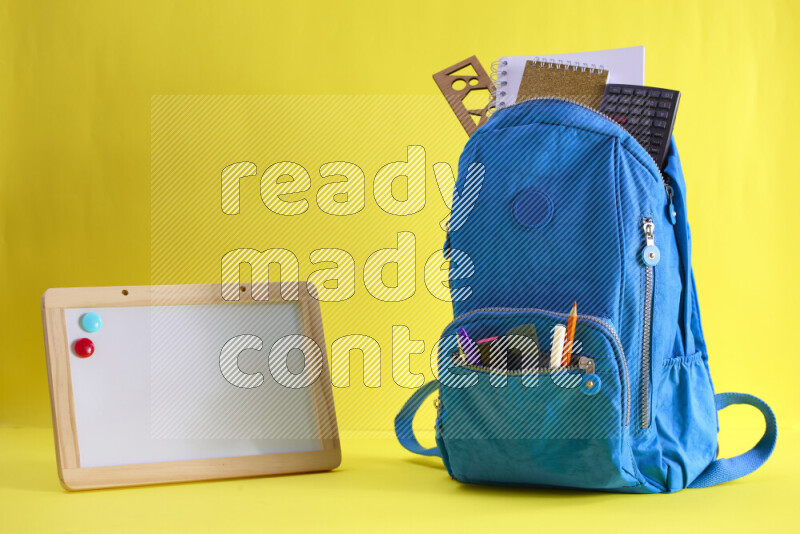 A school bag with assorted school supplies in and beside it on yellow background