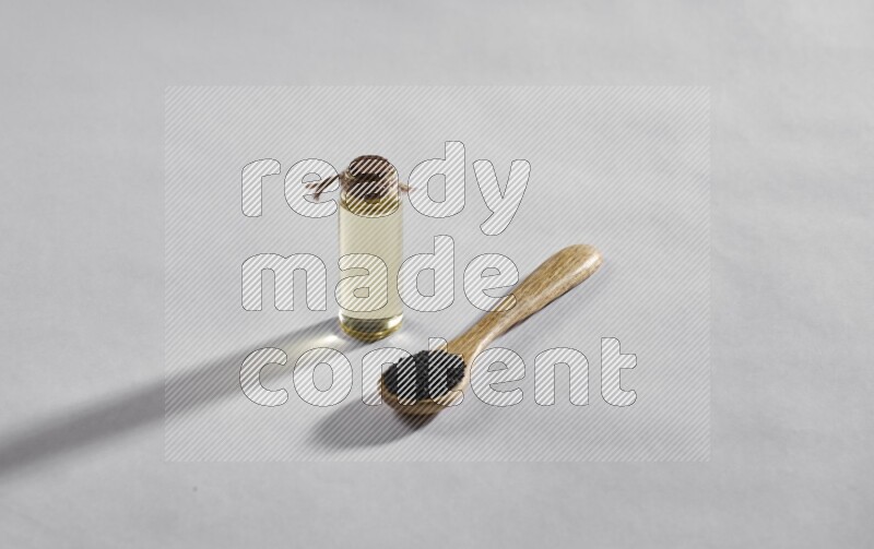 ملعقة خشبية ممتلئة ببذور حبة البركة مع زجاجة من زيت حبة البركة على أرضية بيضاء