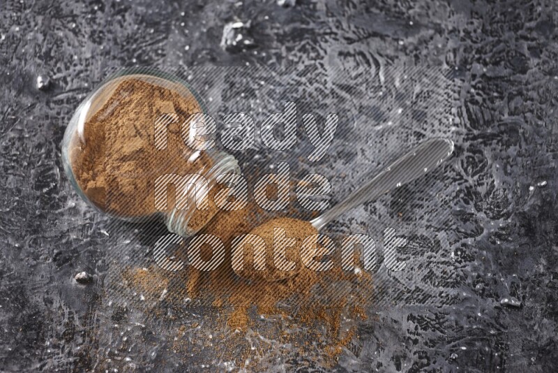 Herbal glass jar full of cinnamon powder flipped and a metal spoon on textured black background