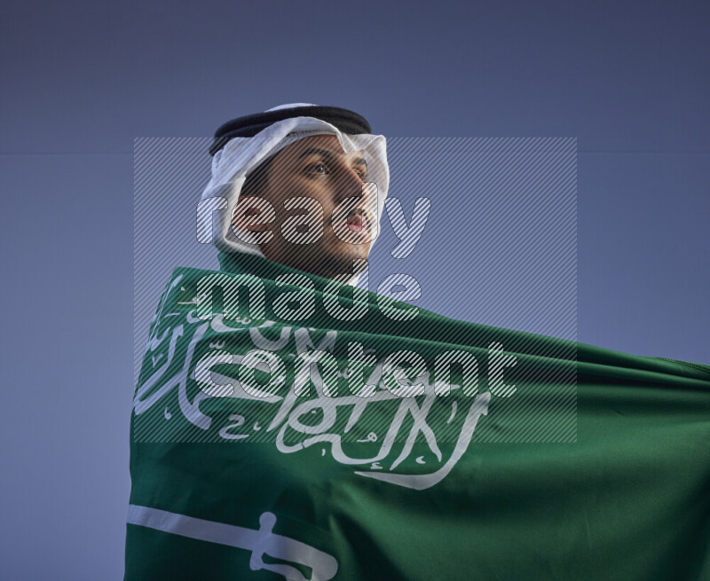 A close-up shot of Saudi man wearing thob and white shomag wrapping big Saudi flag on gray background