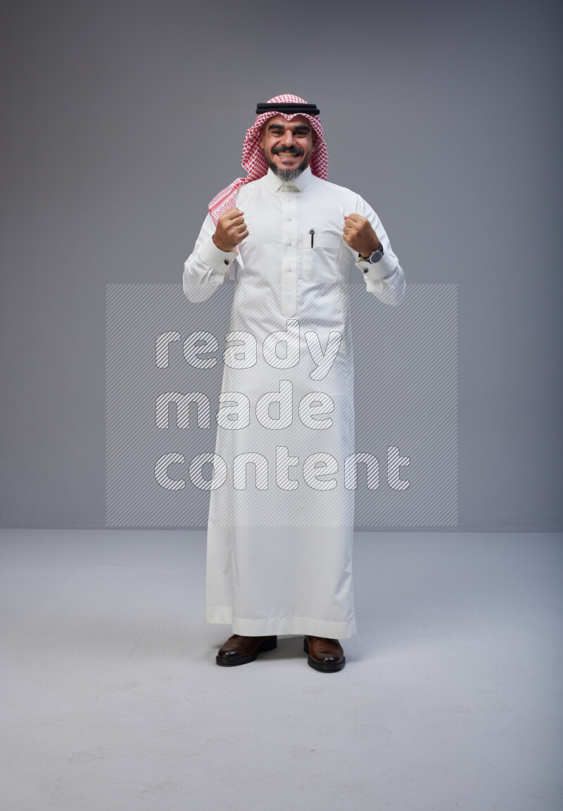 Saudi man Wearing Thob and red white Shomag standing interacting with the camera on Gray background