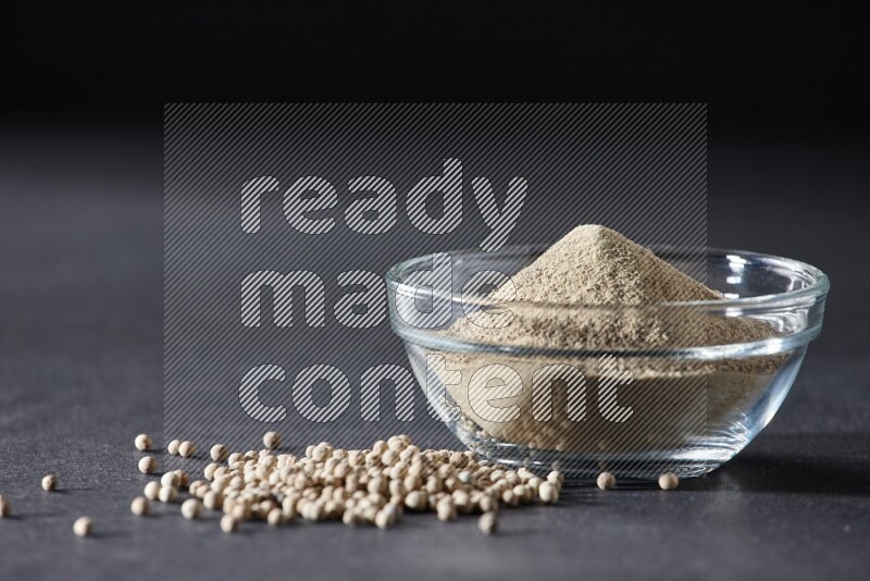 A glass bowl full of white pepper powder with white pepper beads on black flooring