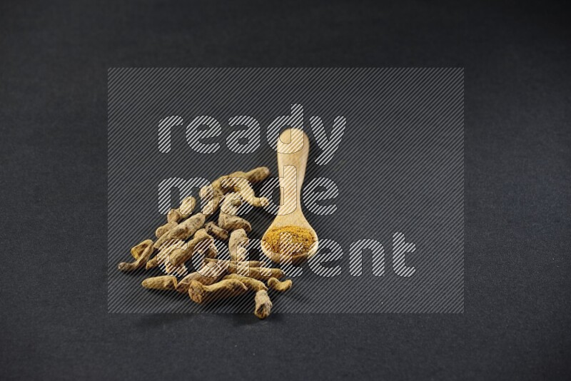 ملعقة خشبية ممتلئة ببودرة الكركم مع أصابع الكركم المجففة على خلفية سوداء