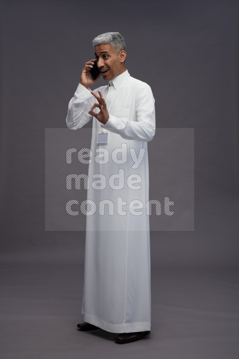 Saudi man wearing thob with neck strap employee badge standing talking on phone on gray background