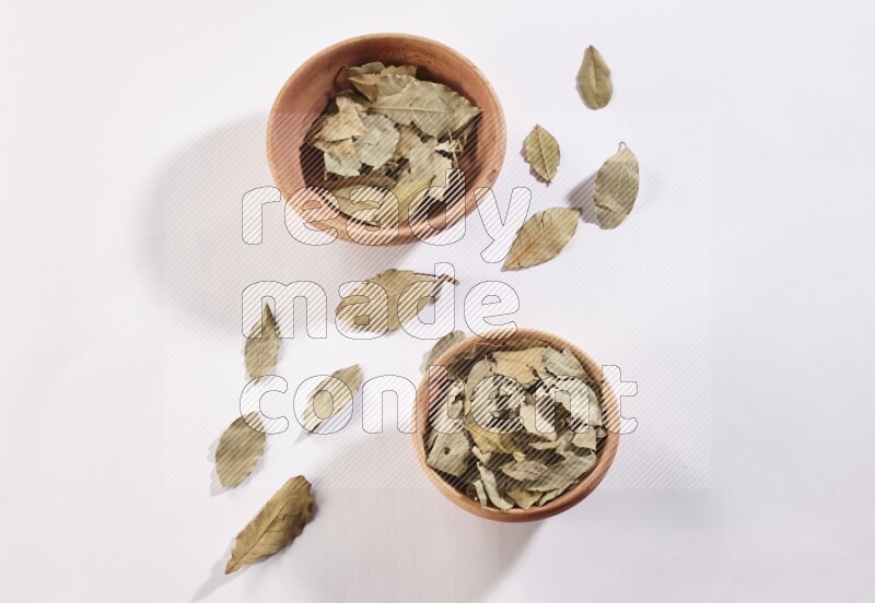 2 wooden bowls full of dried bay leaves with more leaves spread on white flooring