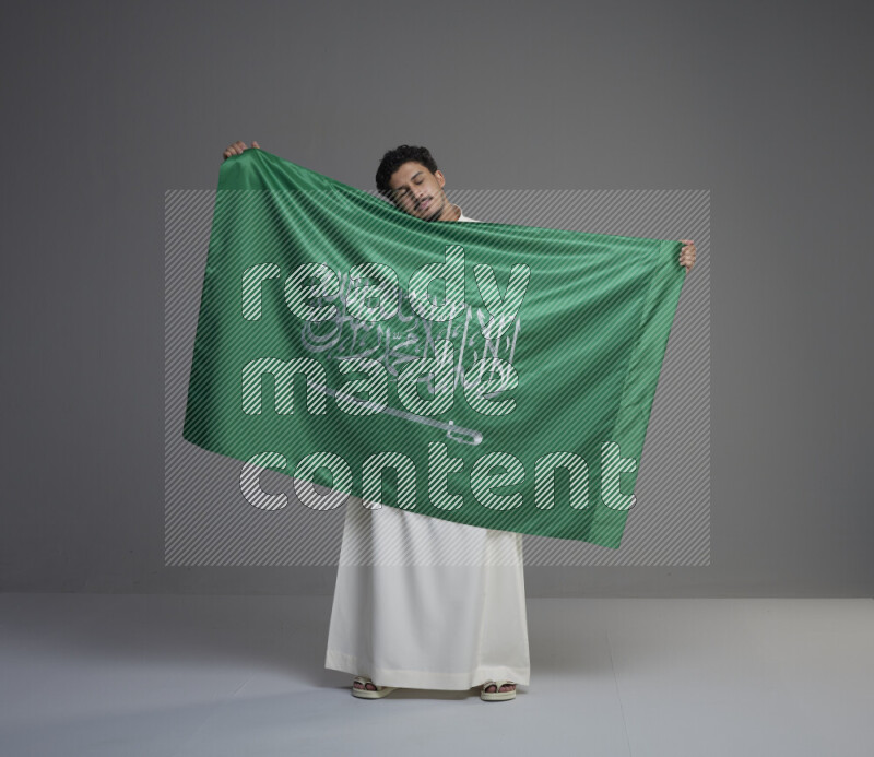 A Saudi man standing wearing thob holding big Saudi flag on gray background