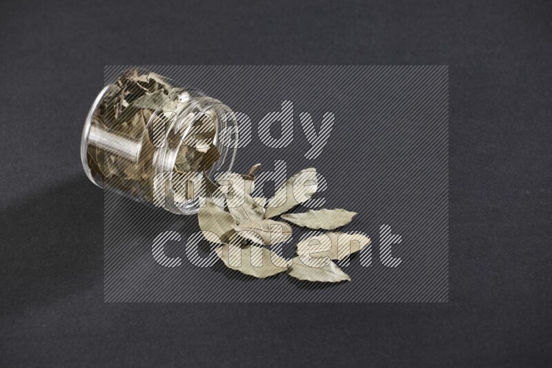 A glass jar full of dried bay leaves and jar is flipped and leaves spread out on black flooring