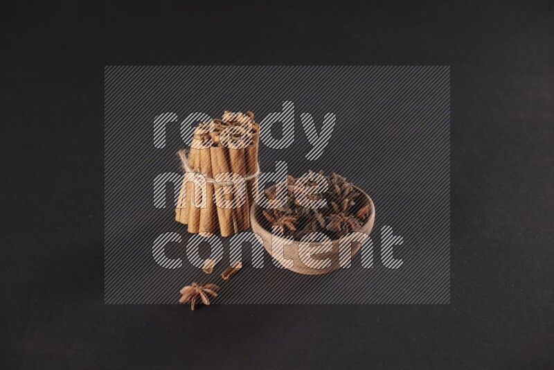 A stacked and bounded cinnamon sticks and a wooden bowl full of star anise on a black background