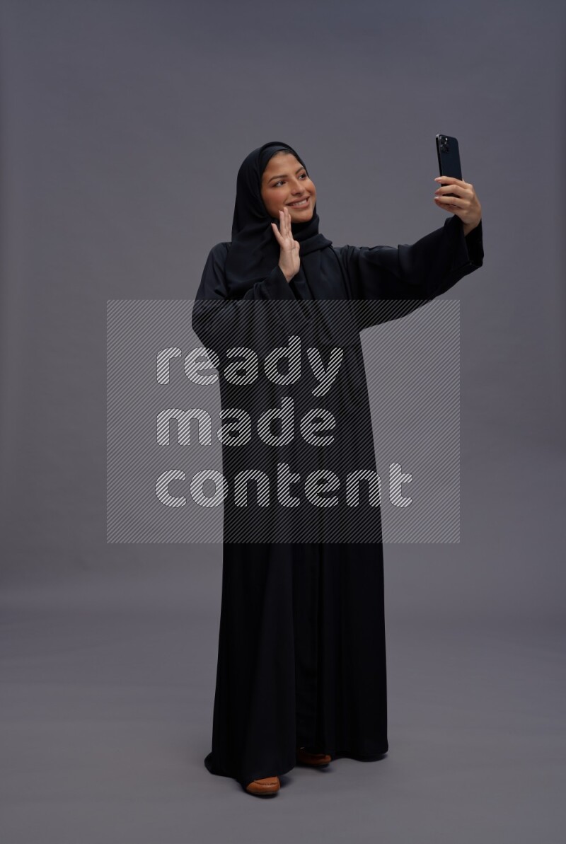 Saudi woman wearing Abaya standing taking selfie on gray background