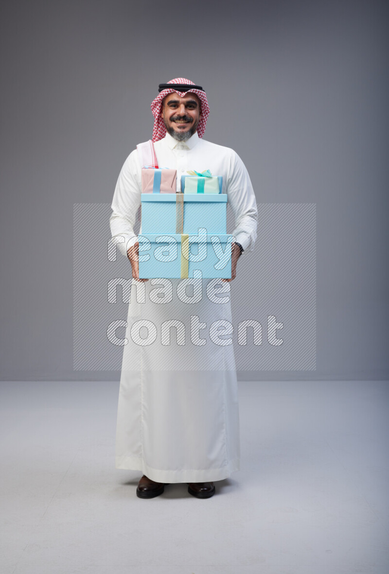 Saudi man Wearing Thob and red Shomag standing holding gift box on Gray background