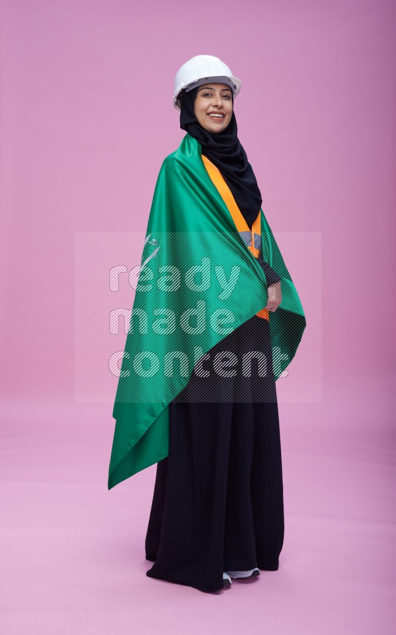 Saudi woman wearing Abaya with engineer vest and helmet standing holding Saudi flag on pink background
