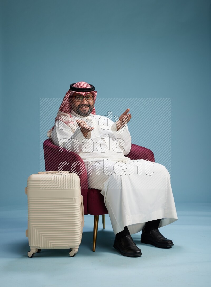 Saudi Man with shimag sitting on chair with a travel luggage on blue background