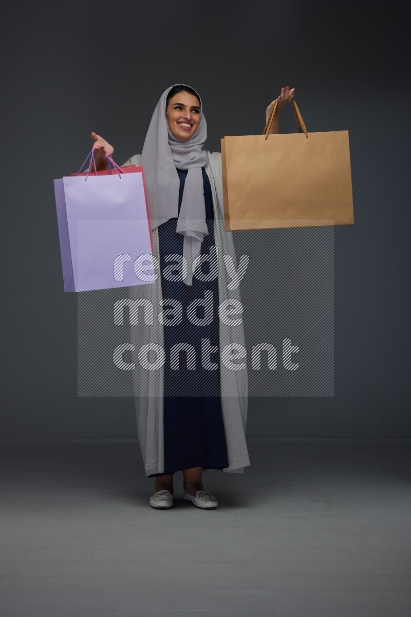 A Saudi woman wearing a light gray Abaya and head scarf standing and holding shopping bags on a grey background