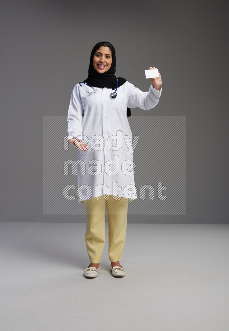 Saudi woman wearing lab coat with stethoscope standing holding ATM card on Gray background