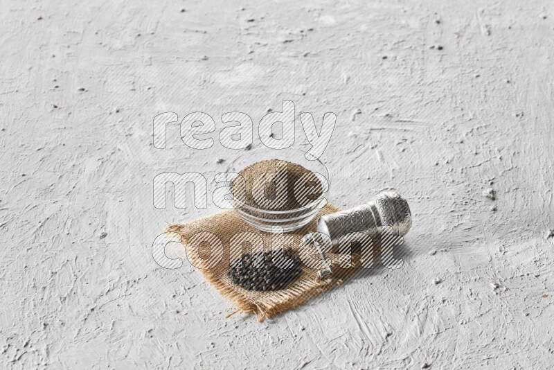 A glass bowl full of black pepper powder, black pepper beads and a turkish metal grinder on burlap fabric on textured white flooring