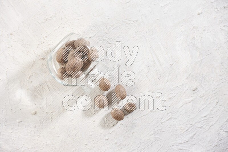 A glass spice jar full of whole nutmeg seeds flipped and the seeds spread out on a textured white flooring