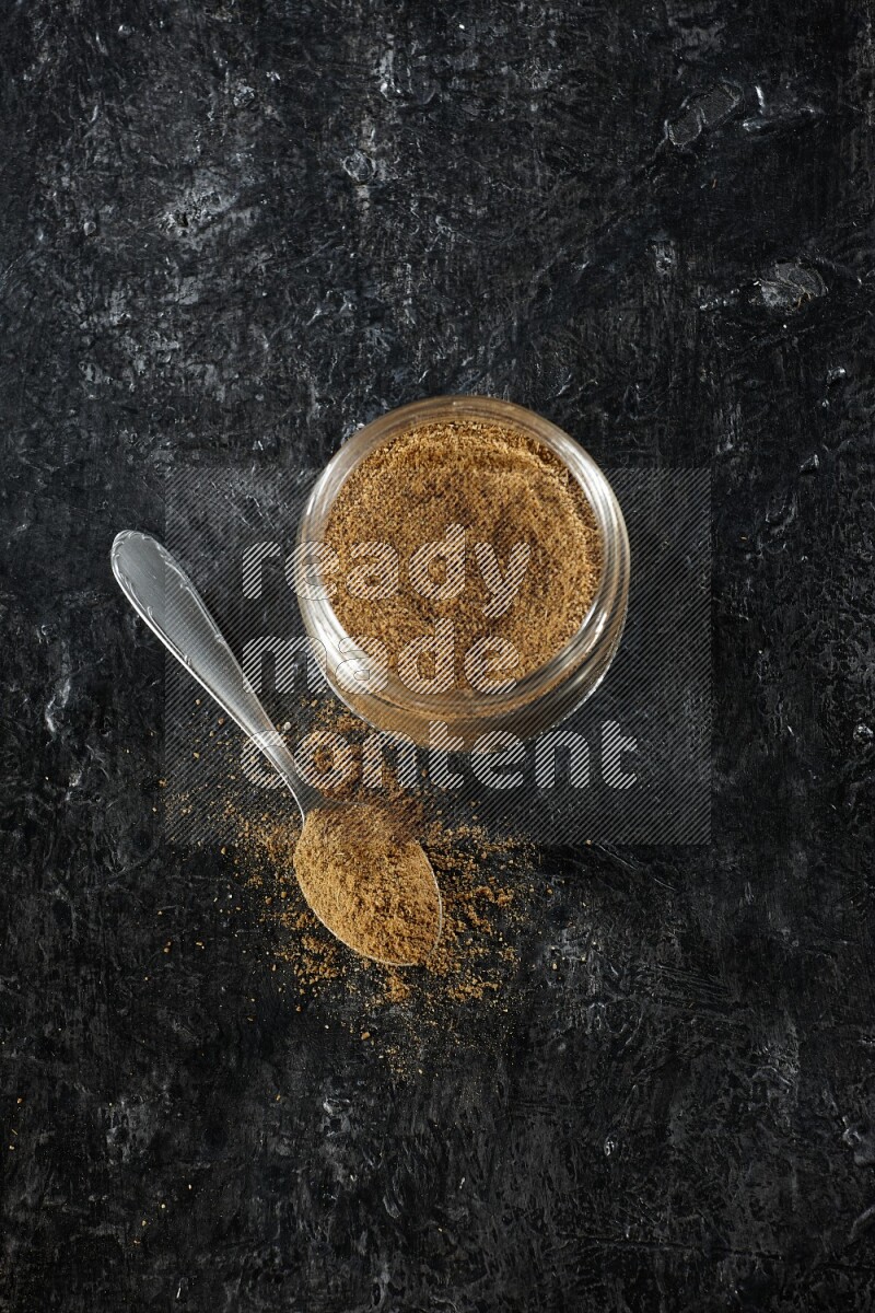 A glass jar and a metal spoon full of cumin powder on a textured black flooring