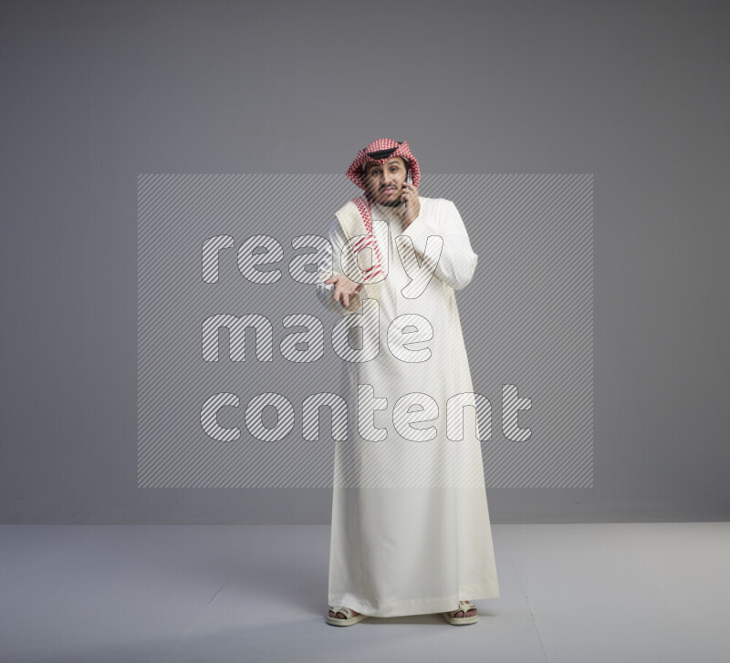 A Saudi man standing wearing thob and red shomag talking on phone on gray background
