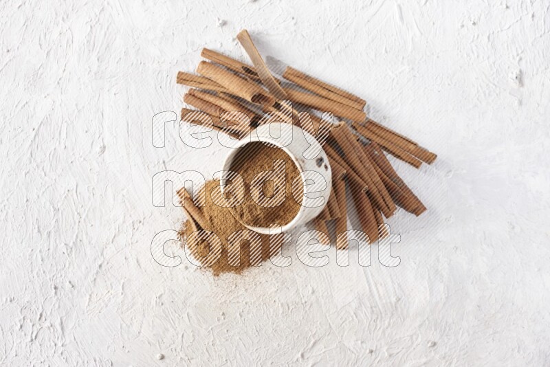 Ceramic beige bowl over filled with cinnamon powder and cinnamon sticks around the bowl on a textured white background in different angles
