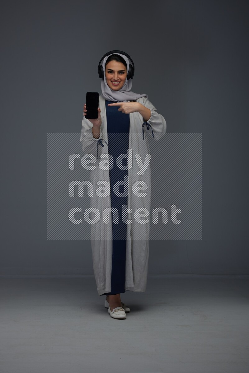 A Saudi woman wearing a light gray Abaya and head scarf setting on a dark grey chair and watching on the tablet eye level on a grey background