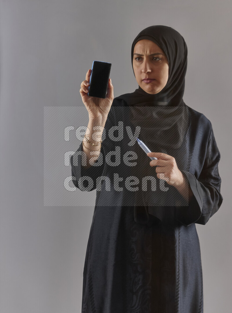 Woman in black abaya and hijab holding a marker pen to write on a transparent board with different reactions and expressions on grey background