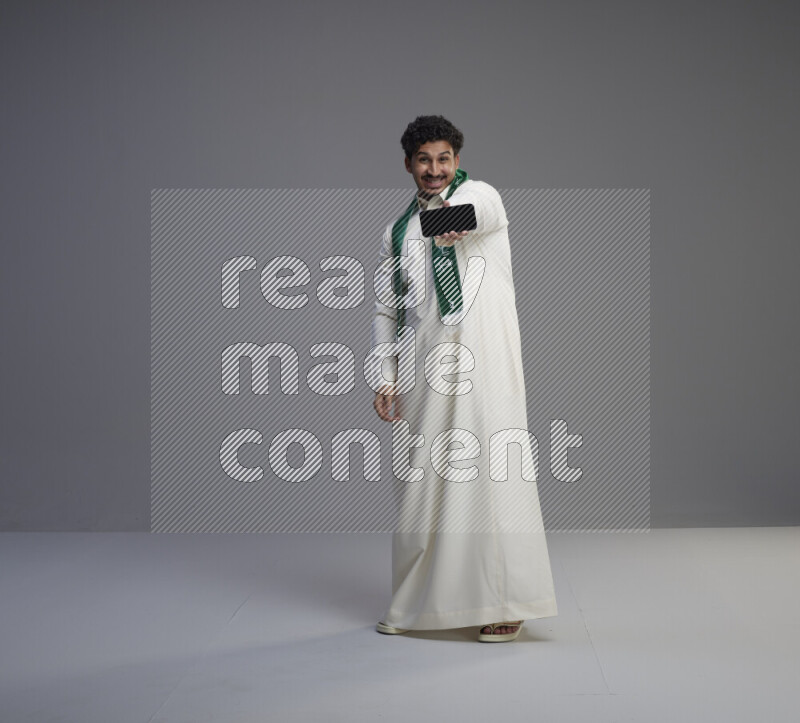 A Saudi man standing wearing thob and Saudi flag scarf showing phone to camera on gray background